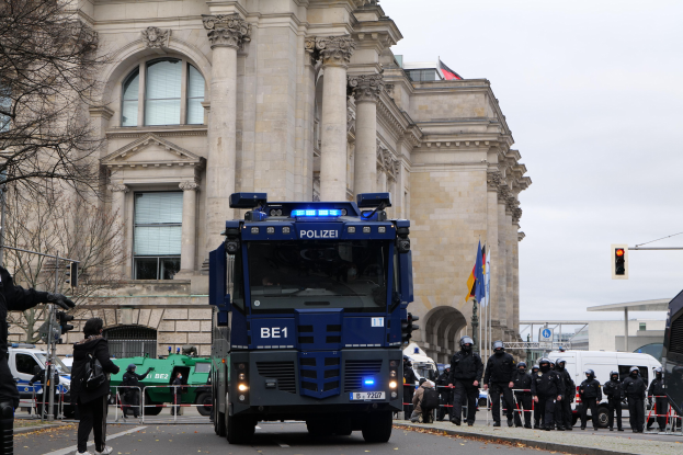 Gruppe von Polizeibeamten vor einem großen Gebäude mit architektonischen Details, Fahrzeugen auf der Straße, einer Person mit einer Kamera links, Bäumen, Verkehrsampeln, Fahnenmasten und einem klaren blauen Himmel.