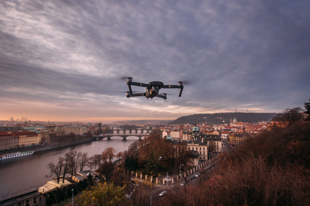 Ein Drohne, die über einer Stadt mit Gebäuden, Bäumen und Fahrzeugen fliegt, mit einem Fluss und einer Brücke im Hintergrund.