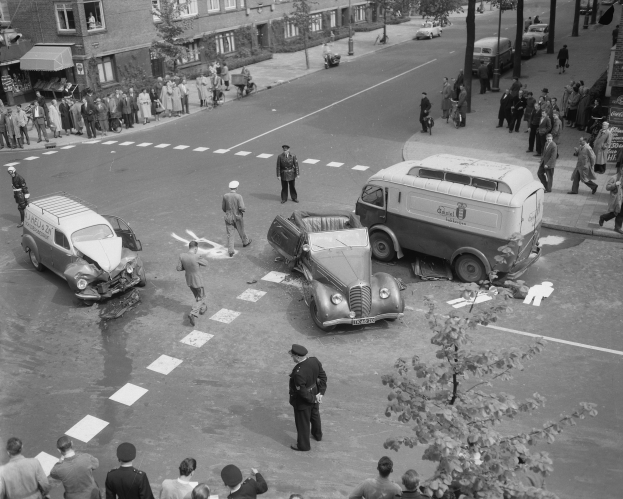 Schwarz-weiß-Szene eines Autounfalls an einer Straßenkreuzung mit mehreren Fahrzeugen, darunter ein Lieferwagen, und einer Gruppe von Schaulustigen; Gebäude, Bäume, Strommasten und Texttafeln sind im Hintergrund zu sehen.