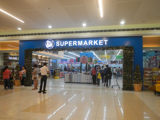 Eine Gruppe von Menschen vor einem Supermarkt, einige mit Einkaufstäschen, mit einem Einkaufswagen, Topfpflanze, Regalen mit Gegenständen, Schildern und einer Decke mit Deckenleuchten.