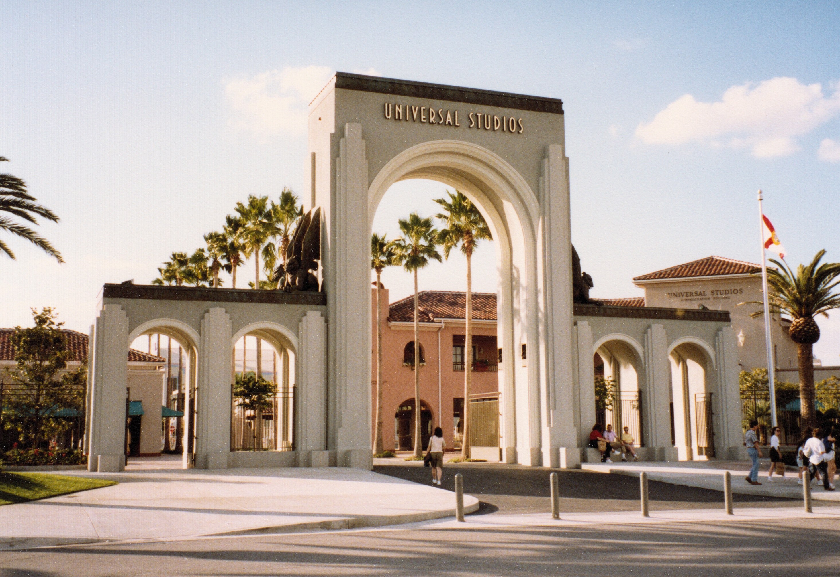 Eingang von Universal Studios Hollywood mit einem Bogen, einer Statue, Menschen, Bäumen, Gebäuden und einem wolkenverhangenen Himmel.