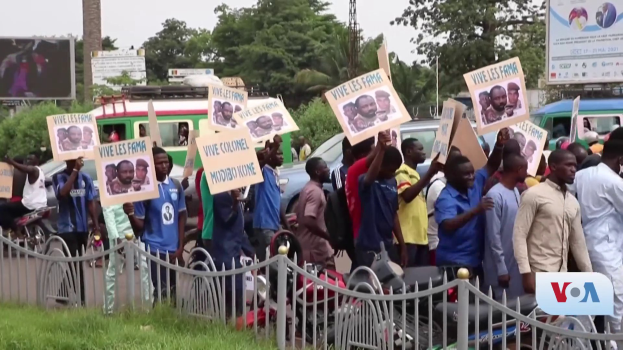 Eine Gruppe von Menschen hält Schilder, wahrscheinlich protestierend, vor einem Zaun mit Fahrzeugen, Bäumen, Pfählen, Absperrungen und einem Bildschirm im Hintergrund, unter einem sichtbaren Himmel mit Gras am unteren Bildrand.