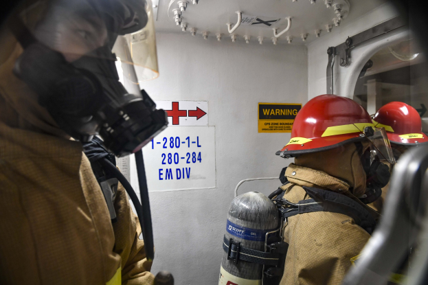 Zwei Feuerwehrleute in Schutzausrüstung stehen nebeneinander in einem Raum mit textgefüllten Tafeln an der Wand, Rohren und Equipment darüber.