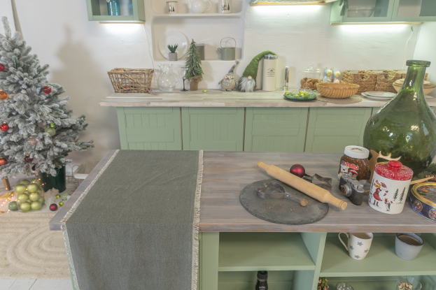 Eine Küche mit grünen Schränken und einem Weihnachtsbaum in der Ecke, mit einem Tisch mit einer Decke, Nudelholz, Flasche, Schüssel, Tassen und anderen Gegenständen, während Körbe, Teller, Schüsseln, Gläser und Spielzeuge auf einer Plattform sind, mit Wänden, die mit Lichtern und Schränken geschmückt sind.