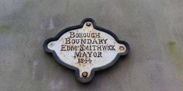 Metallschild an der Seite eines Gebäudes, das die Stadtteilgrenze markiert, mit "Stadtteilgrenze Edm Smithwick Bürgermeister" in fetten weißen Buchstaben auf schwarzem Hintergrund.