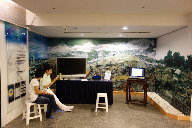 Zwei Frauen sitzen auf Hockern neben einem Tisch mit blauer Decke, einem Fernseher, Laptop, Kalender und einer Flasche; ein weiterer Tisch mit einem Laptop und eine bunte Wand im Hintergrund, mit einem Banner mit Text in der linken Ecke.