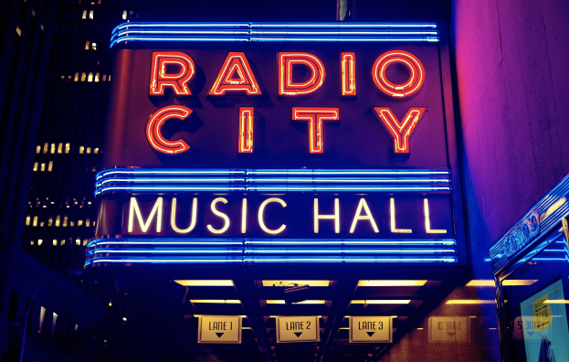 Außenansicht des Radio City Music Hall in New York City bei Nacht, hell erleuchtet mit hohen Gebäuden im Hintergrund, mit einer 'Radio City'-Schrifttafel und zusätzlichen Schildern auf der rechten Seite.