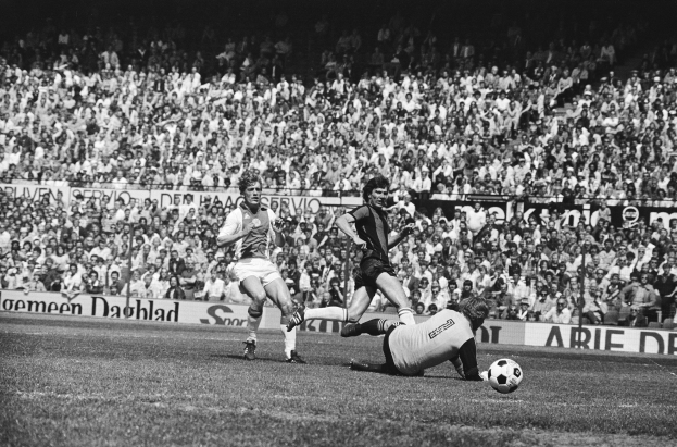 Schwarzes Foto von Männern, die auf einem Feld Füßball spielen, mit Zuschauern im Hintergrund und einer 'Bundesliga 1978-79 Ajax 1-1 Hannover 96'-Tafel.