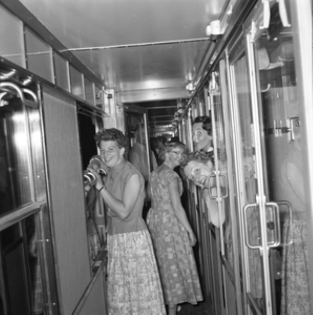 Eine Gruppe von Frauen, die auf einem schwarzen und weißen Zug mit Türen auf beiden Seiten und Lichtern öber ihnen stehen.