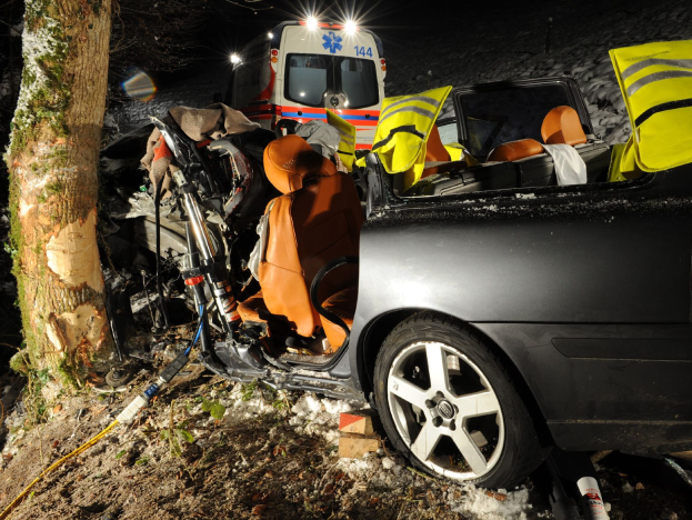 Ein verunfalltes Auto am Straßenrand auf einer schneebedeckten Straße mit einem im Hintergrund sichtbaren Rettungswagen.