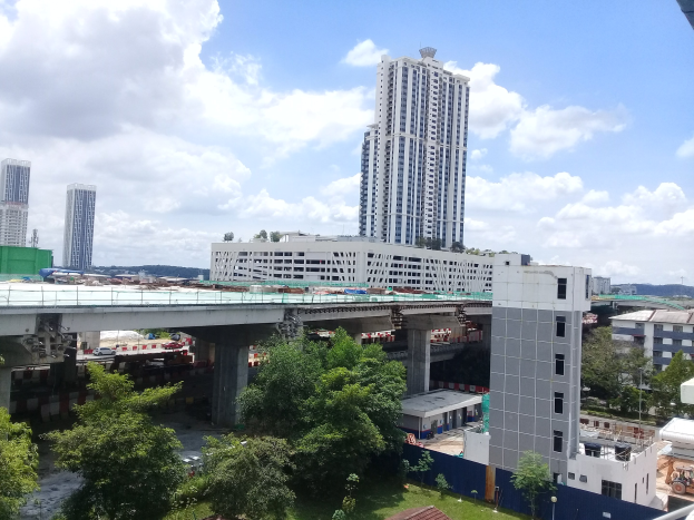 Luftaufnahme einer Stadt mit Gebäuden, Bäumen, Gras, Fahrzeugen, einer Brücke, Masten und einem bewülklichen Himmel, die eine Baustelle für eine neue Wohnanlage zeigt.