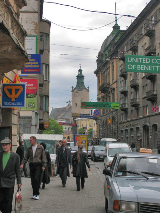 Eine belebte Straßenszene mit Fahrzeugen auf der Straße, Menschen auf einem Fußweg, Gebäuden mit Fenstern, Bäumen, Schildern und anderen Tafeln.
