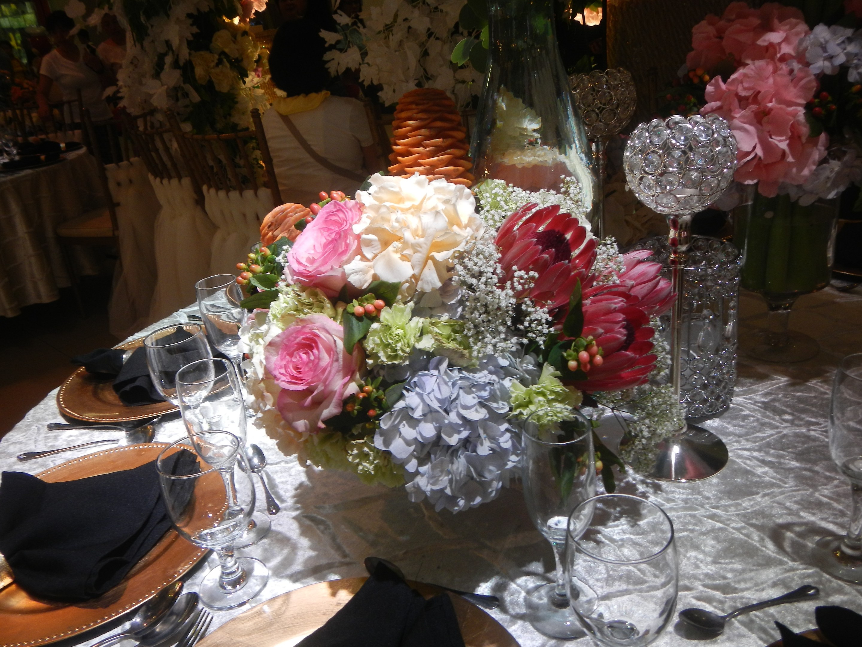 Ein formeller Esstisch mit Tellern, Besteck, Gläsern, Servietten und Blumenvasen, der eine florale Tischdeko und im Hintergrund Stühle und Tische zeigt.
