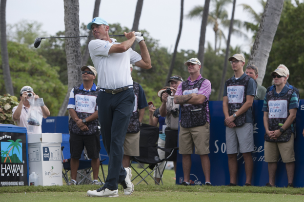 Ein Mann in einem weißen T-Shirt, schwarzen Hosen und einer blauen Mütze schwingt einen Golfschläger auf einem grünen Feld, mit Zuschauern und Kameras hinter ihm unter einem klaren blauen Himmel mit Bäumen.