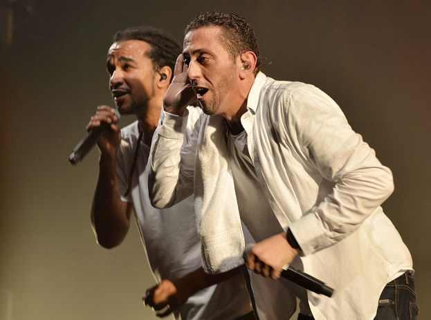 Zwei Männer stehen nebeneinander auf einer Bühne, jeder hält ein Mikrofon in der Hand, tragen Casual-Kleidung während einer Vorstellung.