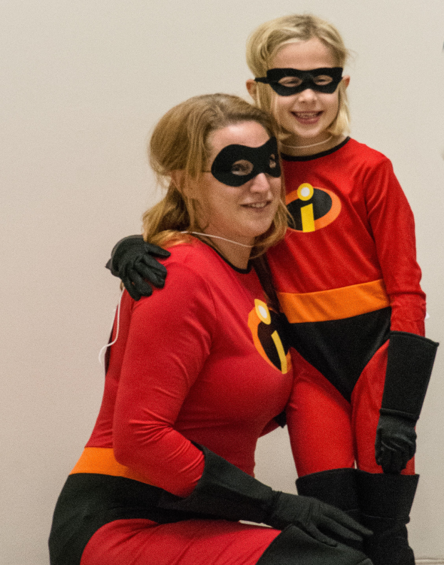 Eine Frau und ein junges Mädchen in roten und schwarzen Kostümen und Masken als die Unglaublichen, wobei die Frau auf dem Boden sitzt und das Mädchen neben ihr steht, vor einer Wand.