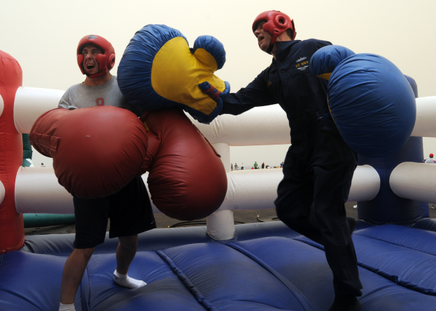 Zwei Männer in Boxhandschuhen und Helmen kämpfen auf einem aufblasbaren Trampolin, mit ein paar Menschen und einem klaren blauen Himmel im Hintergrund.