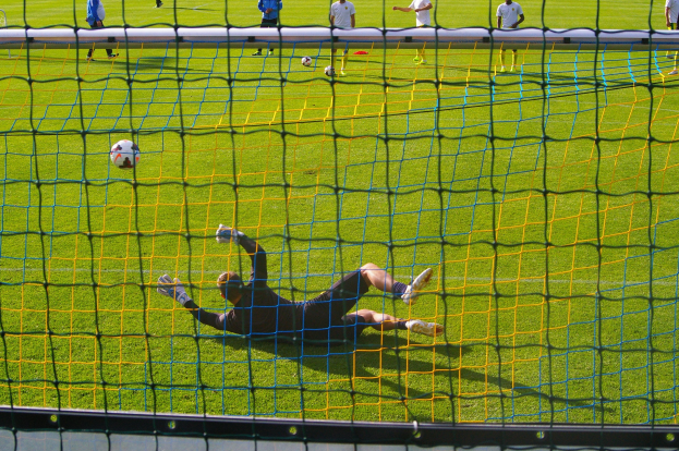 Ein Fußballtorhüter beim Abwehrversuch vor einem Tor, mit einem Netz im Vordergrund und einigen Menschen im Hintergrund, sowie mehreren Fußballen auf dem Boden verteilt.