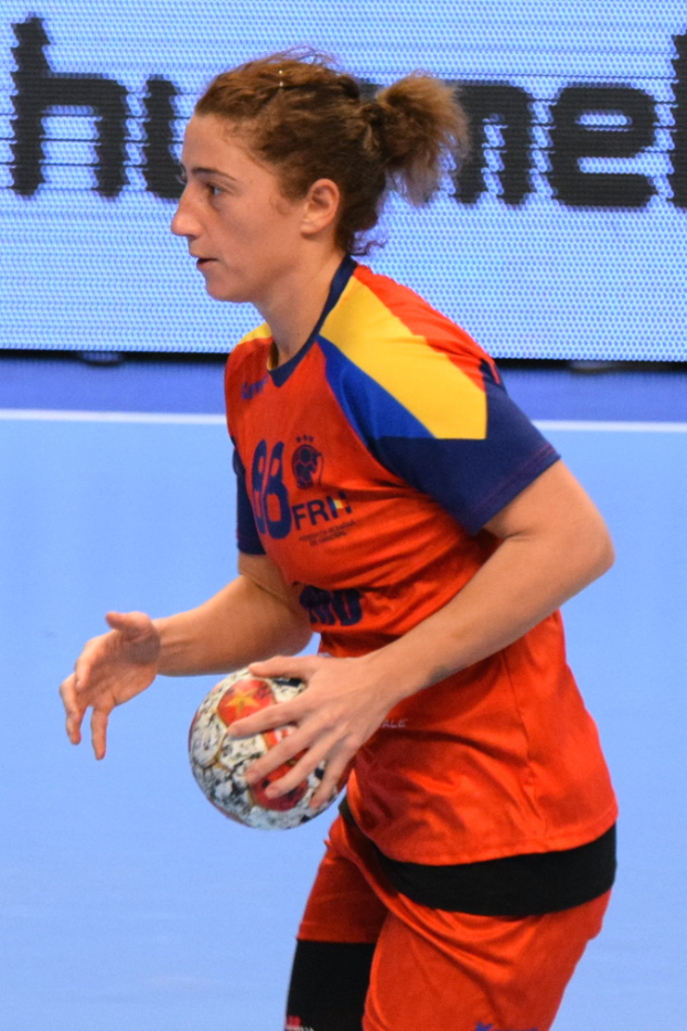 Eine Frau in einem orange-blauen Trikot hält einen Fußball, mit einem Bildschirm im Hintergrund, auf dem Text angezeigt wird.