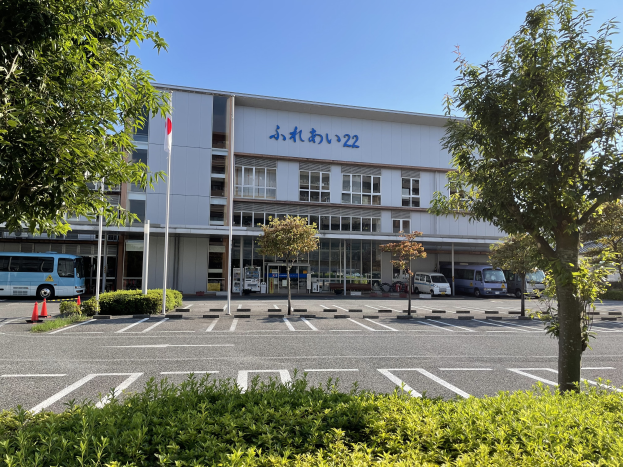 Ein großes Gebäude mit Fenstern und Text, das als Hauptsitz des japanischen Busdienstzentrums dient, mit zahlreichen parkenden Autos davor, umgeben von Pflanzen, Bäumen, Pfählen, Flaggen und Verkehrskegeln, unter einem klaren blauen Himmel.