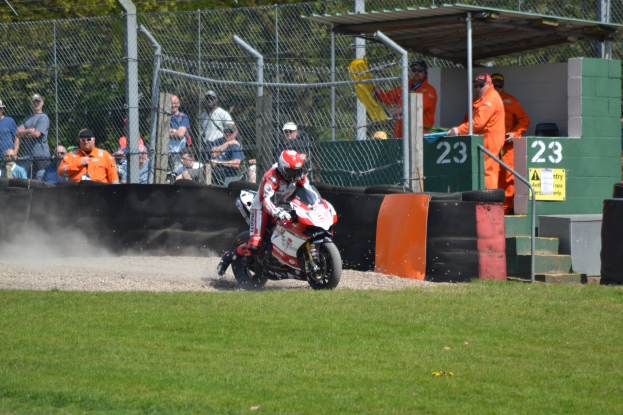 Eine Person in Helm und Handschuhen fährt ein Motorrad über ein grasbewachsenes Feld, mit Zuschauern, Absperrungen, Treppen und einem Schuppen im Hintergrund.