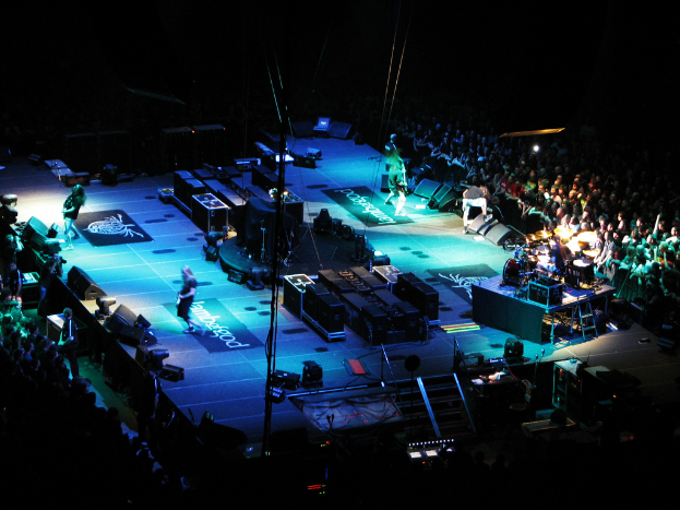 Eine Gruppe von Menschen, die auf einer Bühne mit Lautsprechern, Scheinwerfern und Mikrofonen vor einem großen Publikum spielt.