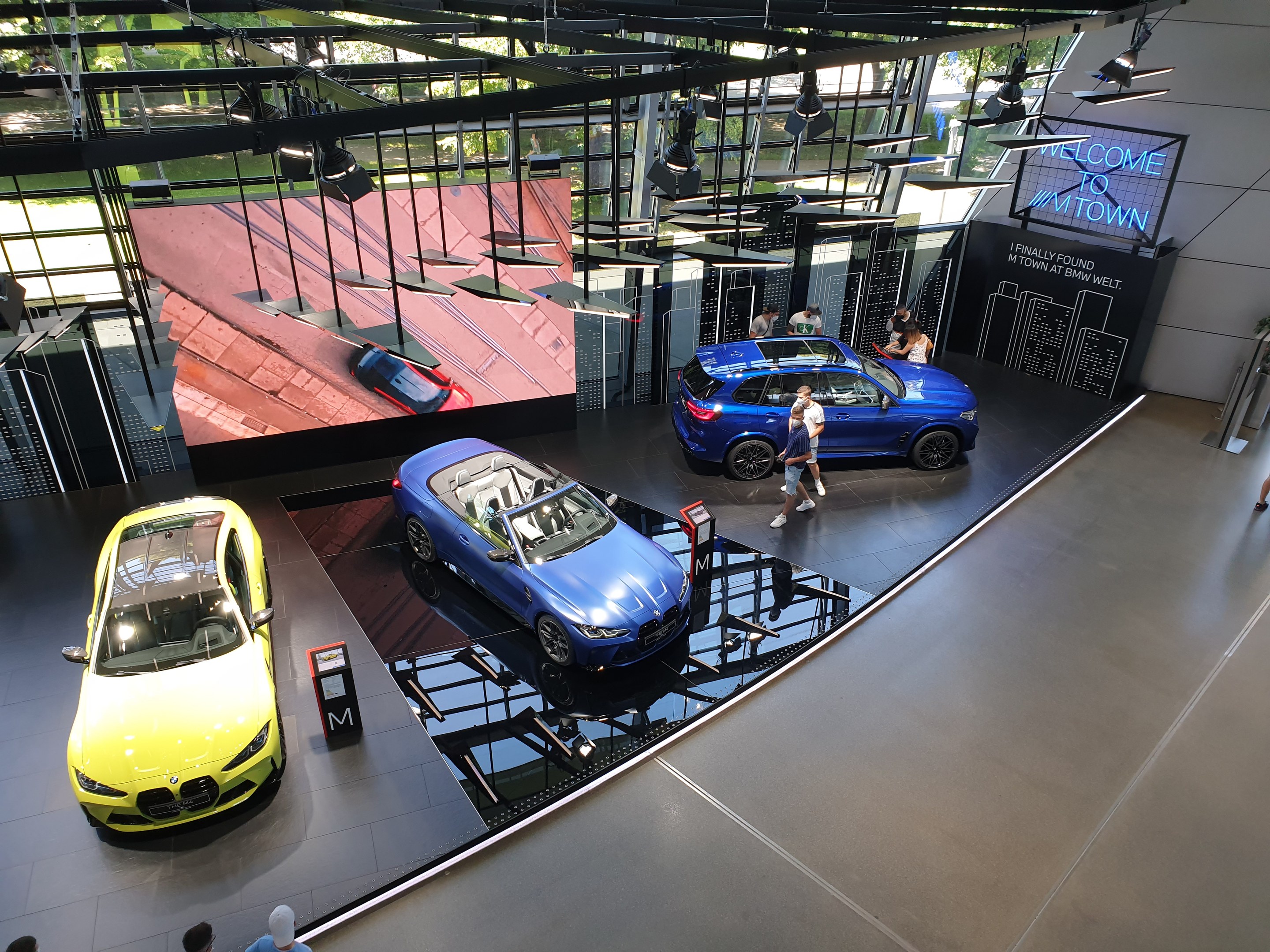 Eine Gruppe von Menschen steht im BMW Museum in München, Deutschland, mit Autos zur Linken und einem Schild mit der Aufschrift "BMW Museum München" zur Rechten, mit Blick auf eine Außenansicht von Bäumen, Gebäuden und Himmel durch Glasfenster.