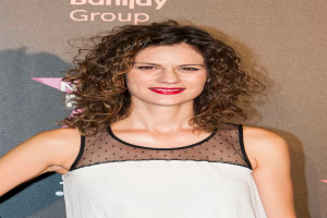 Eine Frau in einem schwarzen Kleid mit tiefem Ausschnitt und schwarzen High Heels, die auf einem roten Teppich steht, strahlt in die Kamera und posiert, mit einem Banner im Hintergrund, auf dem "Maggie Gyllenhaal bei den Billboard Music Awards 2016" steht.