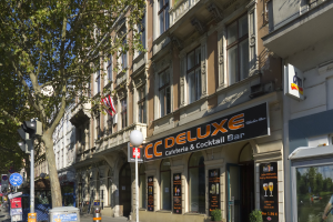 Eine belebte Stadtstraße mit verschiedenen Gebäuden, Fahrzeugen, Fußgängern und Schildern, mit einem hohen Gebäude in der Mitte mit der Aufschrift "CC Deluxe Cafe & Cocktail Bar".