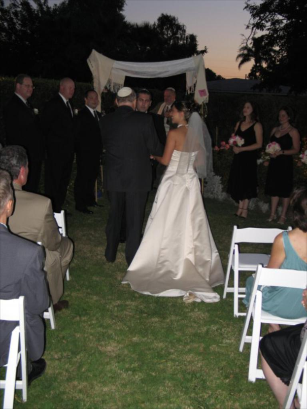 Eine Gruppe von Menschen, die bei einer Hochzeit stehen, wobei Frauen auf der rechten Seite Blumensträuße halten, während andere auf Stühlen sitzen, umgeben von Bäumen.