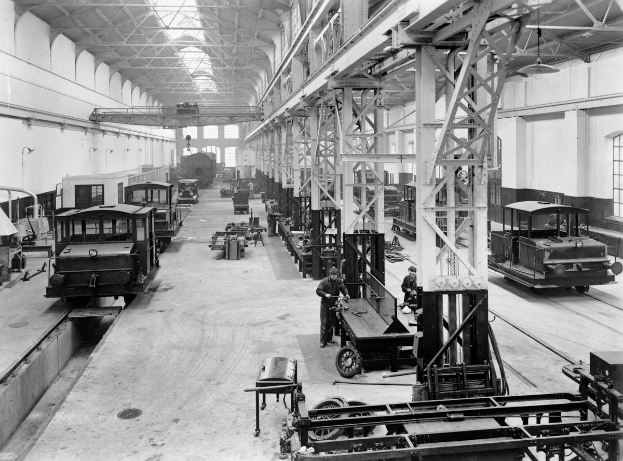 Schwarzes und weißes Foto einer Fabrikhalle mit mehrerenbahnen in Reparatur, Arbeitern, die sich darum kümmern, Metallpfosten, die das Dach stützen, Deckenlampen und Wandfenster.