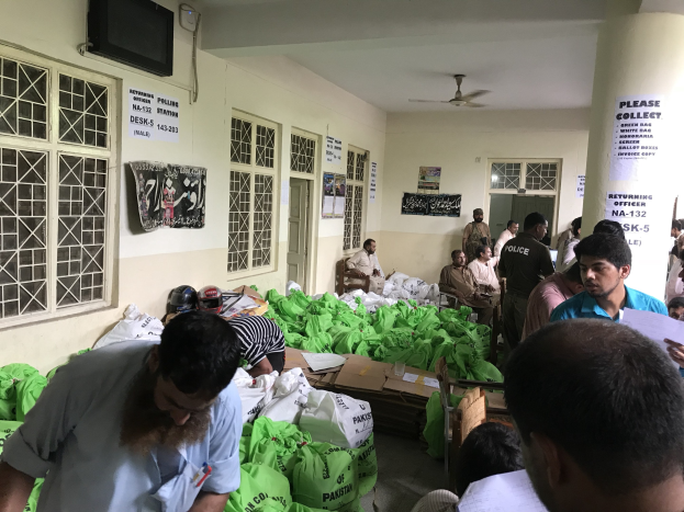 Eine Gruppe von Menschen in einem Raum mit Lebensmittelbeuteln, einige sitzen auf Stühlen und andere stehen, halten Papiere, in der Nähe von Fenstern mit Plakaten, einem montierten Fernseher, Türen und einer Säule, sowie Deckenventilatoren.