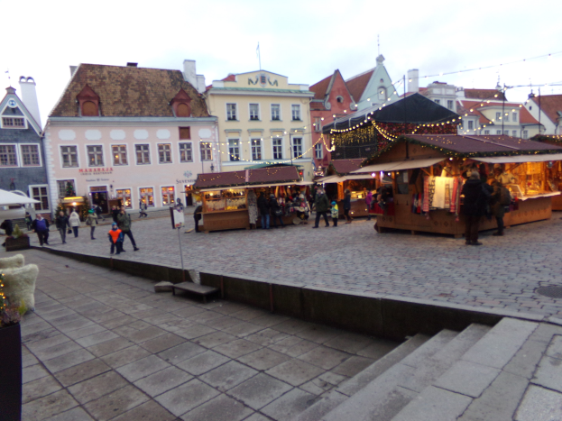 Ein geschäftiger Weihnachtsmarkt in Tallinn, Estland mit Menschen um geschmückte Stände, festliche Gebäude, bewölkter Himmel, Treppe nach oben und Topfpflanzen.
