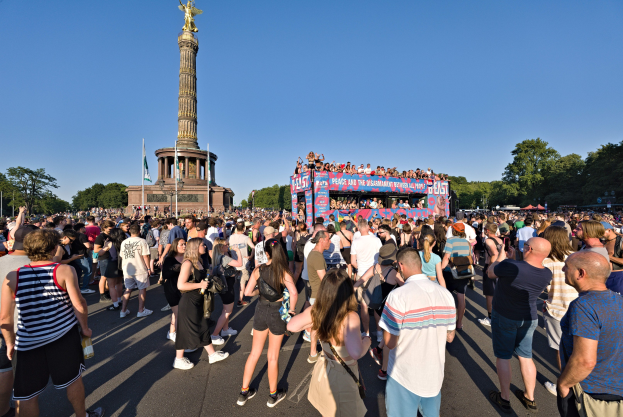 Große Menschenmenge versammelt sich vor einem Denkmal in Berlin mit Fahnen, einem Gebäude mit Säulen und einer Statue, Bäumen und einem klaren blauen Himmel.
