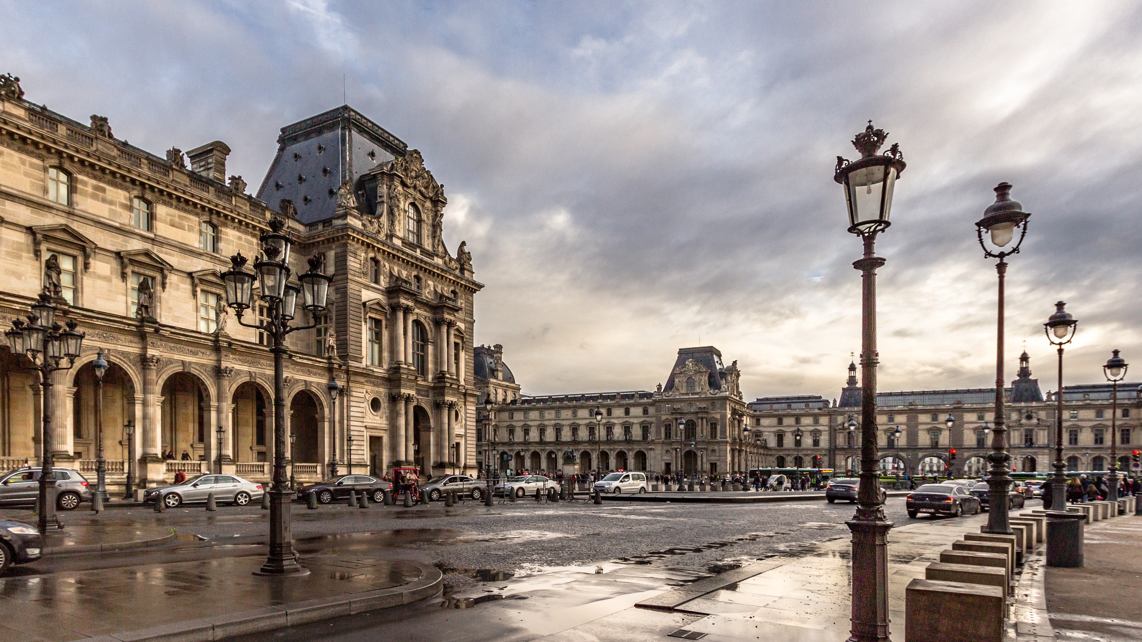 Das Bild zeigt das Louvre-Museum in Paris, Frankreich, mit seinen ikonischen Gebäuden, Straßenlaternen, Straßenlaternen, Kraftfahrzeugen auf der Straße, Menschen auf dem Gehweg und einem Himmel mit Wolken im Hintergrund.