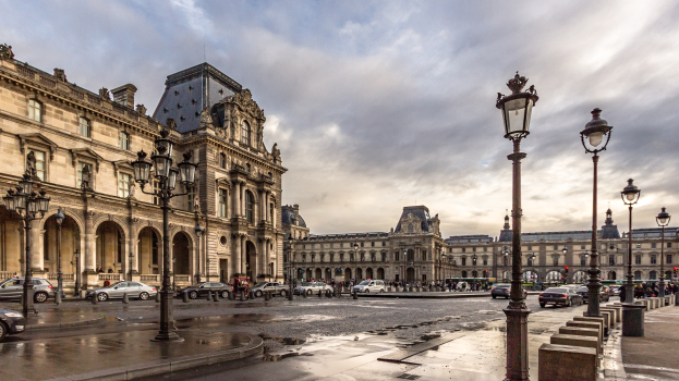 Das Bild zeigt das Louvre-Museum in Paris, Frankreich, mit seinen ikonischen Gebäuden, Straßenlaternen, Straßenlaternen, Kraftfahrzeugen auf der Straße, Menschen auf dem Gehweg und einem Himmel mit Wolken im Hintergrund.