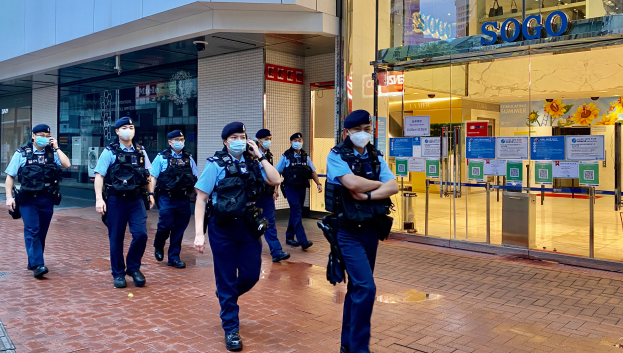 Eine Gruppe von Polizisten in Gesichtsmasken, die vor einem Glasgebäude die Straße entlanggehen, mit einem Geschäft auf der rechten Seite, das Plakate auf seinen Glaswänden hat.