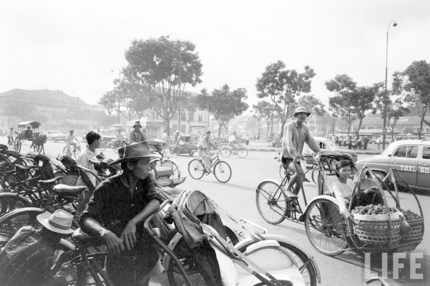 Eine belebte Straße mit Fahrrädern, Autos und anderen Fahrzeugen, mit Bäumen und Gebäuden im Hintergrund.