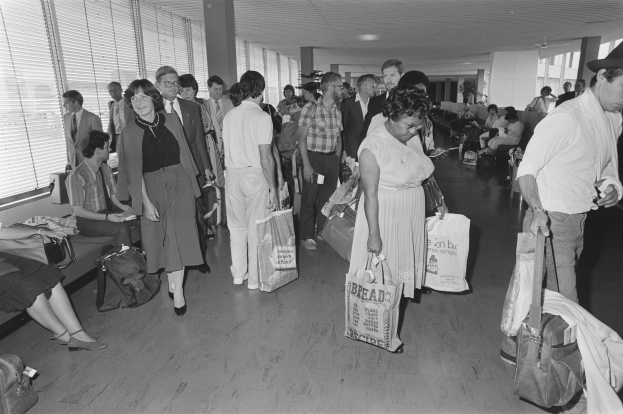 Schwarzes und weißes Foto von Reisenden in einem Flughafen, einige gehen mit Gepäck, andere sitzen auf Bänken oder stehen, mit einem Mann und einer Frau, die nebeneinander stehen und Taschen halten.