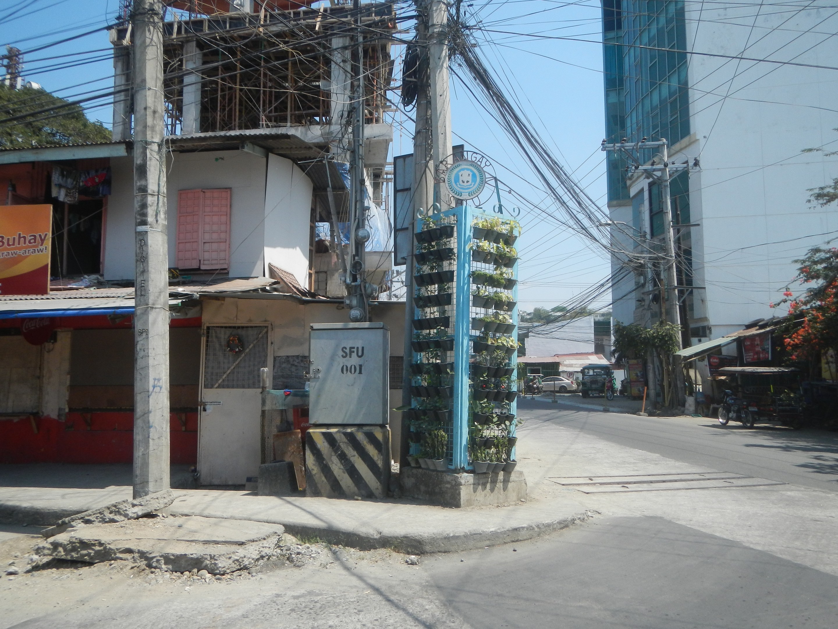 Eine Stadtstraße mit Gebäuden, Strommasten, Kabeln, Namensschildern, Fahrzeugen, Bäumen und einem Himmel im Hintergrund, mit einem Gebäude mit einem Bündel Kabel an der Seite im Vordergrund.