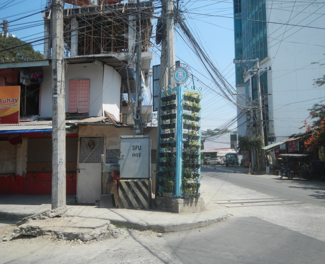 Eine Stadtstraße mit Gebäuden, Strommasten, Kabeln, Namensschildern, Fahrzeugen, Bäumen und einem Himmel im Hintergrund, mit einem Gebäude mit einem Bündel Kabel an der Seite im Vordergrund.