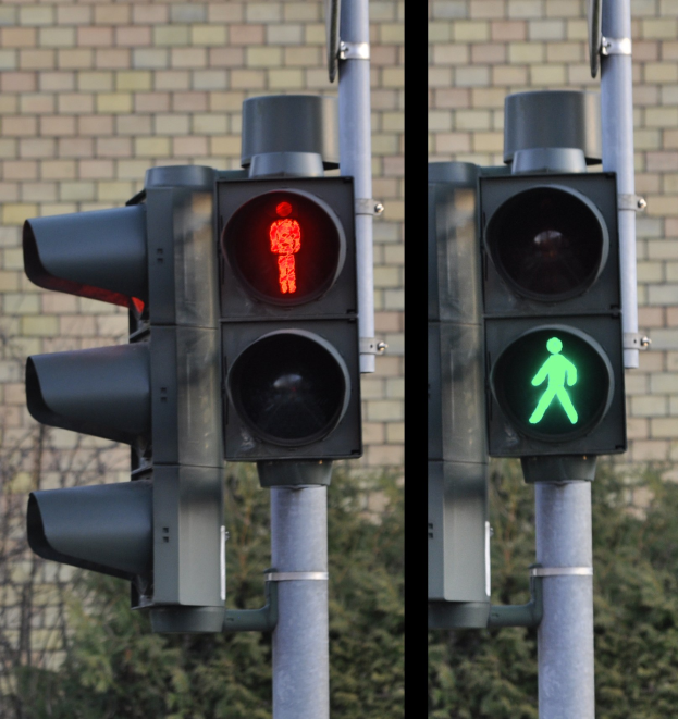 Zwei Ampeln an einem Mast mit roter und grüner Leuchte vor einer Backsteinwand und Bäumen.