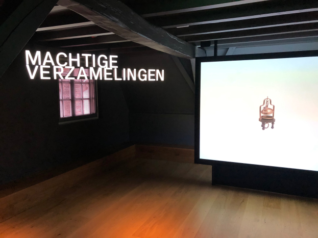 Ausstellungsinstallation 'Machtige Verzamelingen' im Museum of Modern Art in Berlin mit einem großen Bildschirm rechts, Text an der Wand dahinter und einem Fenster links sowie der Decke oben.
