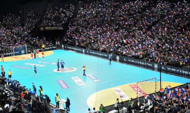 Eine Gruppe von Menschen, die Handball in einem Stadion mit einem Netz auf dem Platz und Zuschauern in den Rängen spielen, während Fotografen die Aktion aufnehmen.