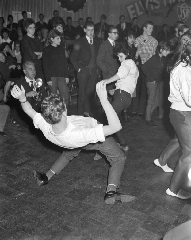 Schwarzes und weißes Bild einer Gruppe von Menschen in Anzügen, die auf einem Boden vor einem Publikum tanzen, mit einer Wand im Hintergrund, auf der etwas geschrieben steht.