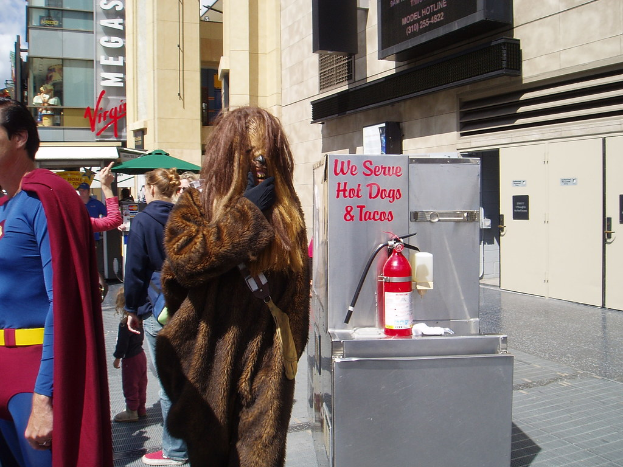 Eine Gruppe von Menschen, einige in Kostümen, versammelte sich vor einem Gebäude mit sichtbaren Schildern und Türen, mit einer Box und einem Feuerlöscher in der Nähe.
