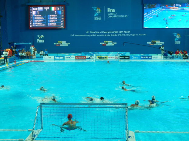 Eine Gruppe von Menschen, die Wasserball in einem großen Pool mit einem Netz in der Mitte spielen, Zuschauer auf der linken Seite und ein Display mit Text und Logos im Hintergrund bei den FINA-Weltmeisterschaften 2016 in Kazan.