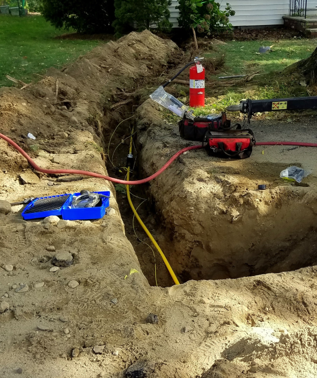 Großes Loch im Boden mit einem Schlauch angeschlossen, umgeben von Werkzeugen einschließlich einem Feuerlöscher und Rohren, in einer Wohngegend mit Bäumen, Pflanzen, Gras und einem Haus im Hintergrund.