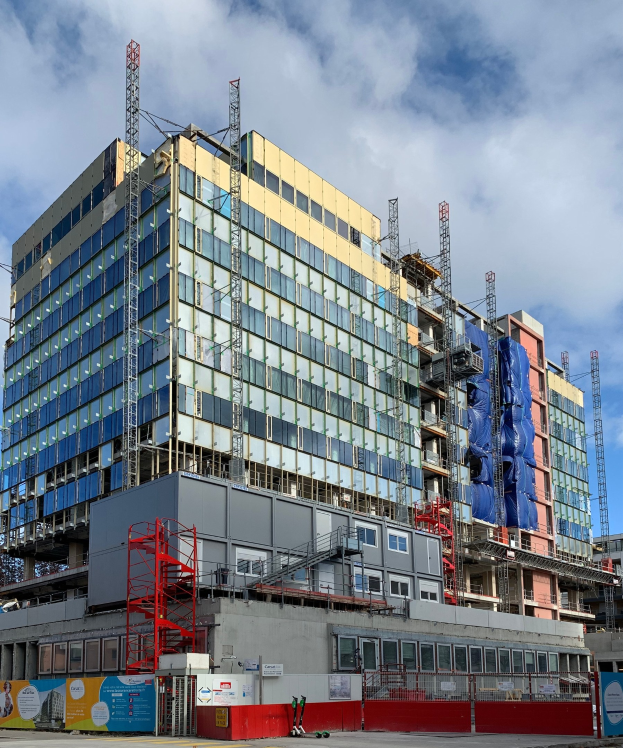 Großes Gebäude im Bau in einer Stadt, mit Glasfenstern, Metallrahmen, einer Treppe, einem Zaun, einem Schild, Pfählen, einem Straßenpfahl, Bäumen und einem bewölkten Himmel.