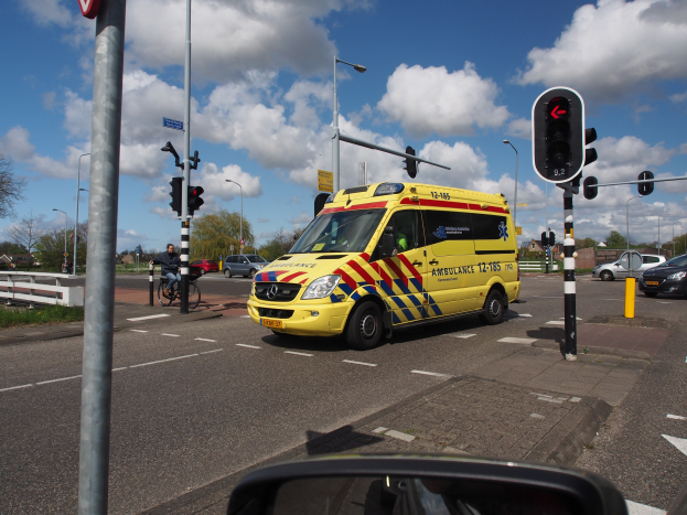 Ambulanz fährt auf einer Straße neben einer Ampel, mit einem Radfahrer im Vordergrund und Laternen, Bäumen, Gebäuden und Lichtmasten im Hintergrund.